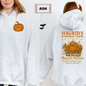 May include: Light gray hoodie with a front pocket and a small orange pumpkin graphic. The back features a design with the text "Hagrid's Pumpkin Farm" and a cart of pumpkins, perfect for Halloween.