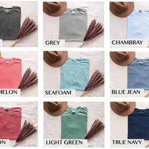 May include: Nine different colored t-shirts, including pepper, grey, chambray, watermelon, seafoam, blue jean, crimson, light green, and true navy. Each shirt is folded and displayed with a brown hat and a bundle of dried flowers.