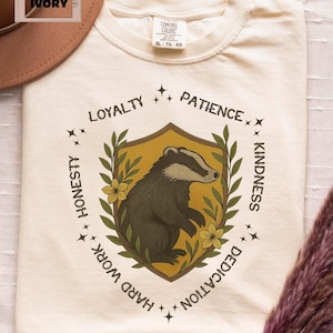 Puede incluir: Camiseta color marfil con un dise&ntilde;o de tej&oacute;n dentro de un escudo, rodeado de texto que incluye "Lealtad" y "Amabilidad". La camiseta es talla XL.