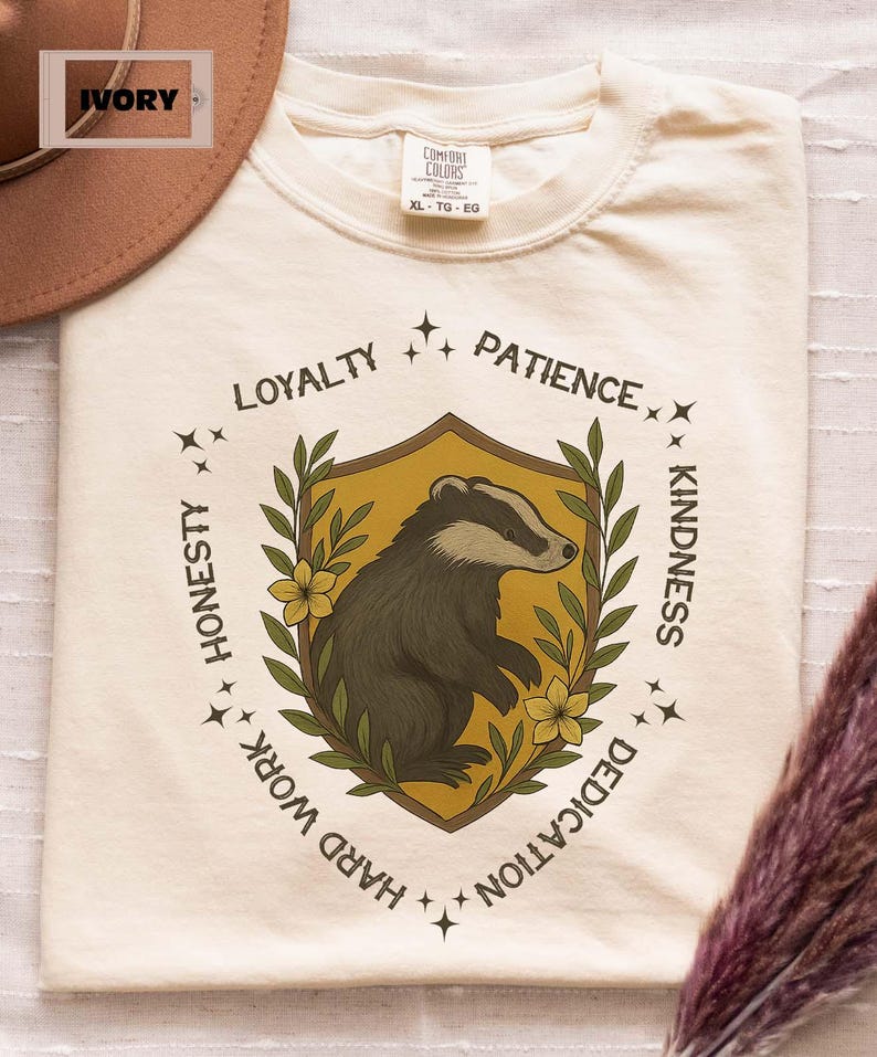 Puede incluir: Camiseta color marfil con un dise&ntilde;o de tej&oacute;n dentro de un escudo, rodeado de texto que incluye "Lealtad" y "Amabilidad". La camiseta es talla XL.