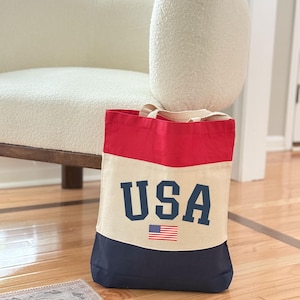 May include: A canvas tote bag with a patriotic design. The top is red, the middle is beige, and the bottom is navy blue. The word "USA" is printed in navy blue block letters, with a small American flag below.