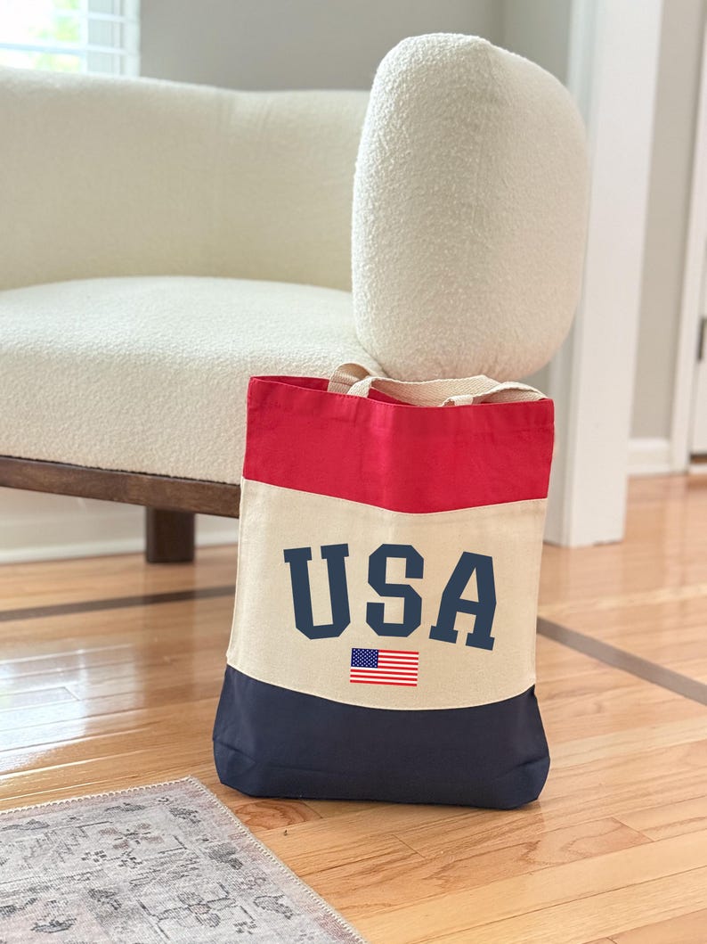 May include: A canvas tote bag with a patriotic design. The top is red, the middle is beige, and the bottom is navy blue. The word "USA" is printed in navy blue block letters, with a small American flag below.