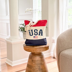 May include: A canvas tote bag with a patriotic design. The top is red, the middle is off-white, and the bottom is navy blue. The word "USA" is printed in navy blue block letters, with a small American flag beneath it. The bag has off-white handles.