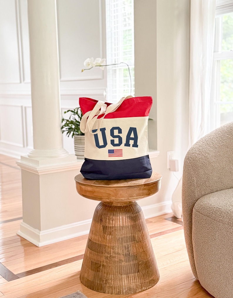 May include: A canvas tote bag with a patriotic design. The top is red, the middle is off-white, and the bottom is navy blue. The word "USA" is printed in navy blue block letters, with a small American flag beneath it. The bag has off-white handles.
