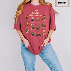 Woman wearing a  crimson t-shirt featuring a holiday design titled “The 12 Christmas Cats of Wizard School,” showing cute illustrated cats dressed as magical characters. Festive Christmas decor in the background including tree, pillows, and seasonal accents.