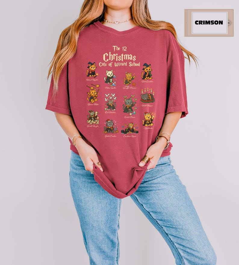 Woman wearing a  crimson t-shirt featuring a holiday design titled “The 12 Christmas Cats of Wizard School,” showing cute illustrated cats dressed as magical characters. Festive Christmas decor in the background including tree, pillows, and seasonal accents.