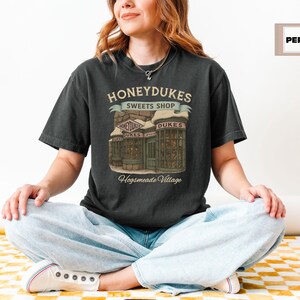 Op de afbeelding: Een zwart T-shirt met een afbeelding van Honeydukes Sweets Shop uit de Harry Potter-serie. De afbeelding toont een vintage-stijl illustratie van de buitenkant van de winkel met de tekst "Honeydukes Sweets Shop" en "Hogsmeade Village".