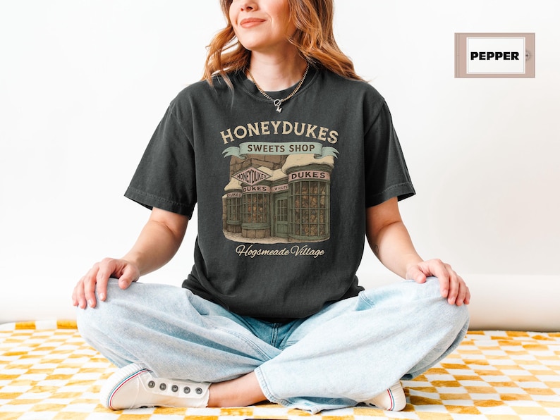 Op de afbeelding: Een zwart T-shirt met een afbeelding van Honeydukes Sweets Shop uit de Harry Potter-serie. De afbeelding toont een vintage-stijl illustratie van de buitenkant van de winkel met de tekst "Honeydukes Sweets Shop" en "Hogsmeade Village".