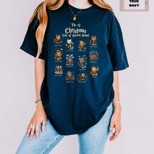 Woman wearing a true navy t-shirt featuring a holiday design titled “The 12 Christmas Cats of Wizard School,” showing cute illustrated cats dressed as magical characters. Festive Christmas decor in the background including tree, pillows, and seasonal accents.