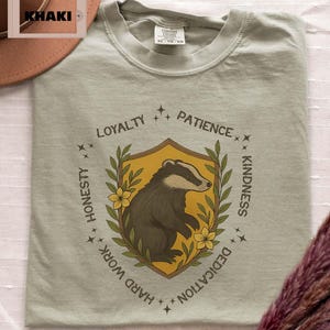 Puede incluir: Camiseta caqui con un escudo que representa un tej&oacute;n, rodeado de palabras como 'Lealtad' y 'Bondad'. El tej&oacute;n es marr&oacute;n y blanco sobre un fondo amarillo. La camiseta tiene un ajuste c&oacute;modo.