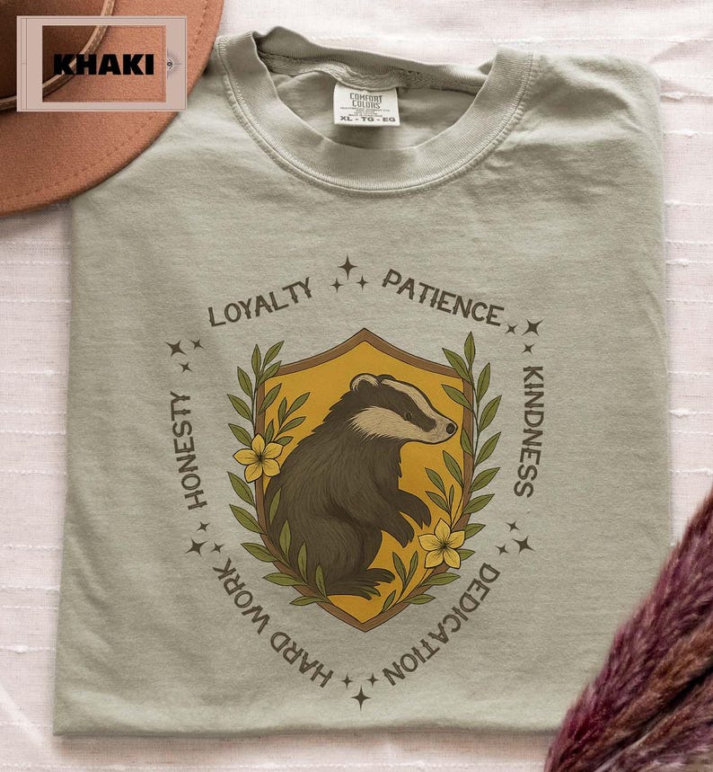 Puede incluir: Camiseta caqui con un escudo que representa un tej&oacute;n, rodeado de palabras como 'Lealtad' y 'Bondad'. El tej&oacute;n es marr&oacute;n y blanco sobre un fondo amarillo. La camiseta tiene un ajuste c&oacute;modo.