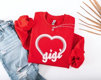 May include: Red sweatshirt with a white heart and the word "gigi" in pink inside the heart. The sweatshirt is folded and laid on a white surface with a pair of blue jeans.