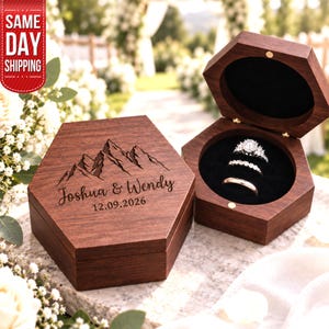 May include: A hexagonal dark wood ring box with a hinged lid, featuring an engraved mountain range, the names Joshua & Wendy, and the date 12.09.2026. The open box interior is lined with black velvet and holds three silver-toned rings.