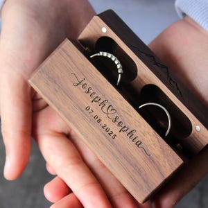 May include: A wooden ring box with two compartments for rings. The box is engraved with the names "Joseph & Sophia" and the date "07.08.2025".