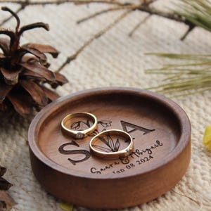 Puede incluir: Un plato redondo de madera para anillos con las letras S y A grabadas, una ilustraci&oacute;n floral y el texto "Saara & Abigail Jan 08 2024". Dos anillos dorados con peque&ntilde;as piedras transparentes descansan dentro de la bandeja circular.