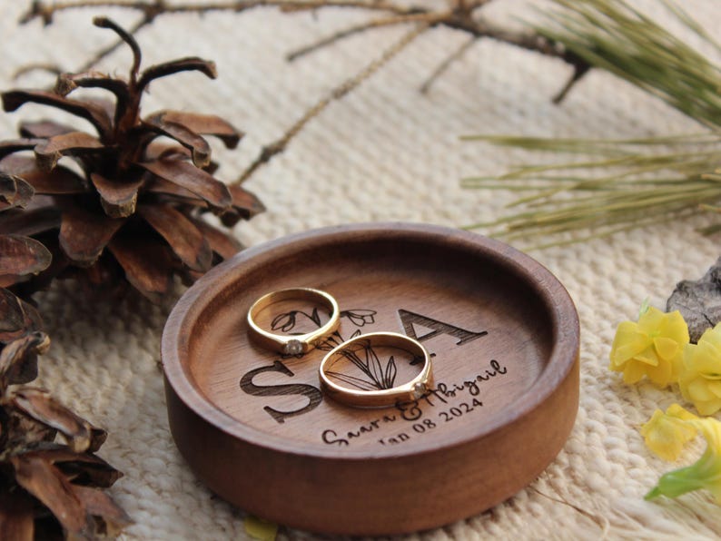 Puede incluir: Un plato redondo de madera para anillos con las letras S y A grabadas, una ilustraci&oacute;n floral y el texto "Saara & Abigail Jan 08 2024". Dos anillos dorados con peque&ntilde;as piedras transparentes descansan dentro de la bandeja circular.