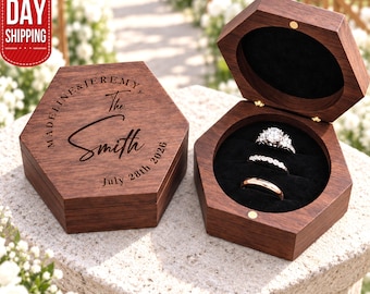 May include: A hexagonal dark wood ring box with a hinged lid. The lid is engraved with the text "MADELINE & JEREMY, The Smith, July 28th 2026". The open box features a black velvet interior holding three rings: one diamond ring, one band with small stones, and one plain gold band.
