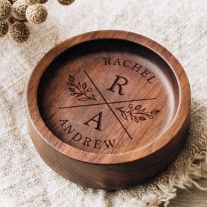 Puede incluir: Un cuenco redondo de madera marr&oacute;n oscuro con un dise&ntilde;o grabado a l&aacute;ser. El centro muestra las letras R y A separadas por l&iacute;neas cruzadas, con los nombres RACHEL y ANDREW grabados a lo largo de los bordes curvos y ramas florales.