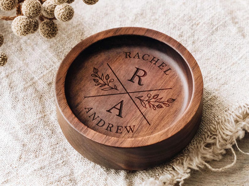Puede incluir: Un cuenco redondo de madera marr&oacute;n oscuro con un dise&ntilde;o grabado a l&aacute;ser. El centro muestra las letras R y A separadas por l&iacute;neas cruzadas, con los nombres RACHEL y ANDREW grabados a lo largo de los bordes curvos y ramas florales.