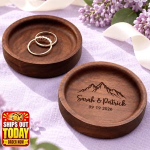 May include: Two round dark brown wooden ring dishes. One dish is empty, while the other holds two silver rings. The second dish features a laser-engraved mountain graphic above the text Sarah & Patrick and the date 09 19 2026.