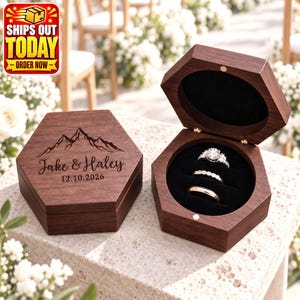 May include: A hexagonal dark wood ring box with a hinged lid. The lid features an engraved mountain graphic, the names Jake & Haley, and the date 12.10.2026. The open box interior is lined with black velvet and holds three rings.
