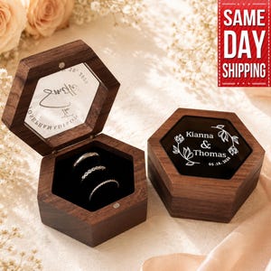 Könnte beinhalten: Zwei sechseckige Holz-Ringboxen, eine offen mit Ringen, die andere geschlossen mit "Kianna & Thomas 05.16.2026" und floralem Design. Die Boxen sind dunkelbraun mit schwarzem Innenraum und Deckel.