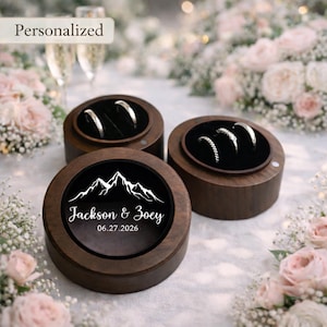 May include: Three round wooden ring boxes with black velvet interiors. Two boxes hold silver rings. One box features a mountain graphic and the names "Jackson & Zoey" with the date "06.27.2026". The word "Personalized" is in the top left corner.