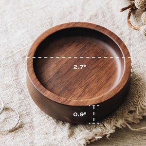 Puede incluir: Un cuenco redondo de madera de color marr&oacute;n oscuro con un acabado liso. El cuenco presenta una veta visible y mide 6,86 cm de di&aacute;metro y 2,29 cm de altura. Dos anillos finos de plata est&aacute;n situados a la izquierda del cuenco de madera.