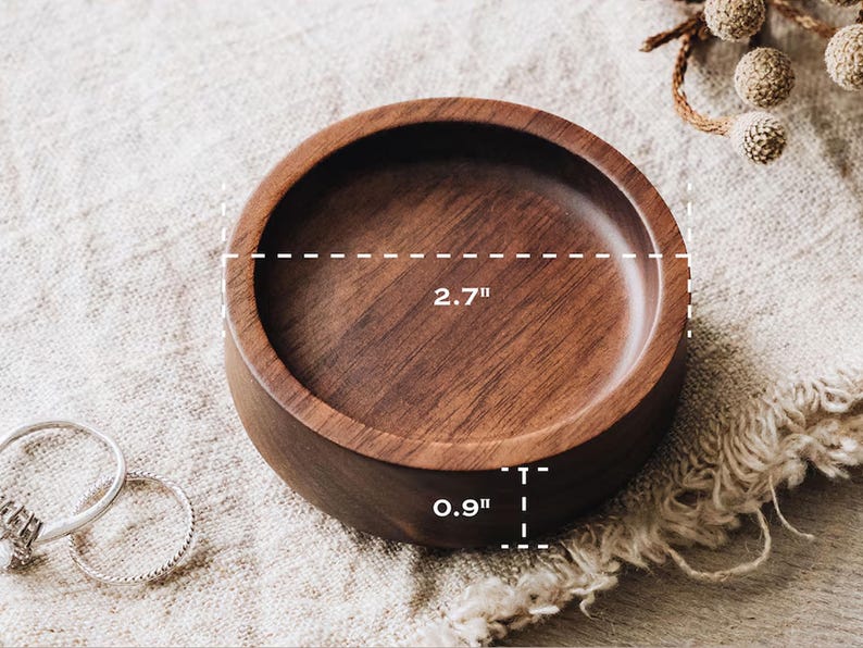 Puede incluir: Un cuenco redondo de madera de color marr&oacute;n oscuro con un acabado liso. El cuenco presenta una veta visible y mide 6,86 cm de di&aacute;metro y 2,29 cm de altura. Dos anillos finos de plata est&aacute;n situados a la izquierda del cuenco de madera.