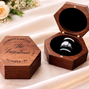 May include: Hexagonal wooden ring box with a dark finish, open to reveal two rings on a black velvet interior. The lid is engraved with names and a date. The closed box is also visible.