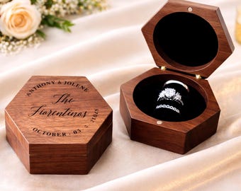 Puede incluir: Caja hexagonal de anillos de madera con acabado oscuro, abierta para revelar dos anillos sobre un interior de terciopelo negro. La tapa está grabada con nombres y una fecha. La caja cerrada también es visible.