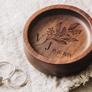 Puede incluir: Un plato redondo de madera marr&oacute;n oscuro con un dise&ntilde;o grabado de rama bot&aacute;nica, las letras V y J, y la fecha 11.20.2024. Dos anillos de tono plateado, uno con una piedra central y otro con una banda trenzada, se sit&uacute;an junto al plato.