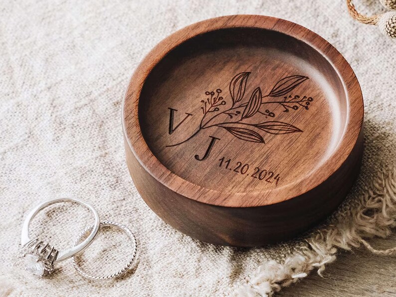 Puede incluir: Un plato redondo de madera marr&oacute;n oscuro con un dise&ntilde;o grabado de rama bot&aacute;nica, las letras V y J, y la fecha 11.20.2024. Dos anillos de tono plateado, uno con una piedra central y otro con una banda trenzada, se sit&uacute;an junto al plato.