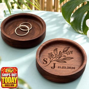 Puede incluir: Dos platos redondos de madera de color marr&oacute;n oscuro para anillos. Uno es liso y contiene dos anillos dorados. El otro presenta un dise&ntilde;o grabado con l&aacute;ser de una rama con hojas, las letras S y J, y la fecha 23.11.2026 en la base.