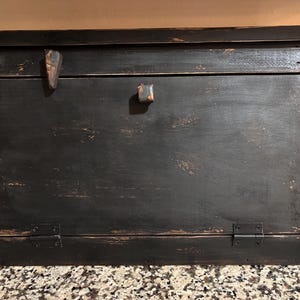 May include: A rectangular wooden bread box with a distressed black finish, featuring a hinged front panel, a small central wooden knob, a decorative wooden accent piece on the upper left, and two black metal hinges at the bottom edge.