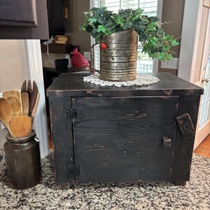 May include: A black wooden cabinet with a distressed finish, featuring a single door with metal hinges and a small latch. On top sits a metallic flour sifter containing green artificial ivy, next to a dark ceramic crock holding wooden kitchen utensils.