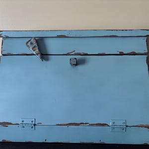 May include: A light blue wooden bread box with a distressed finish showing brown wood underneath. It features a slanted front door with a small square knob, a top lid with a triangular latch, and two metal hinges at the bottom of the front panel.