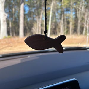 May include: A wooden fish-shaped car air freshener hanging from a black string. The fish is a dark brown color and has a simple, stylized design. The background shows a blurred view of trees and a blue sky.