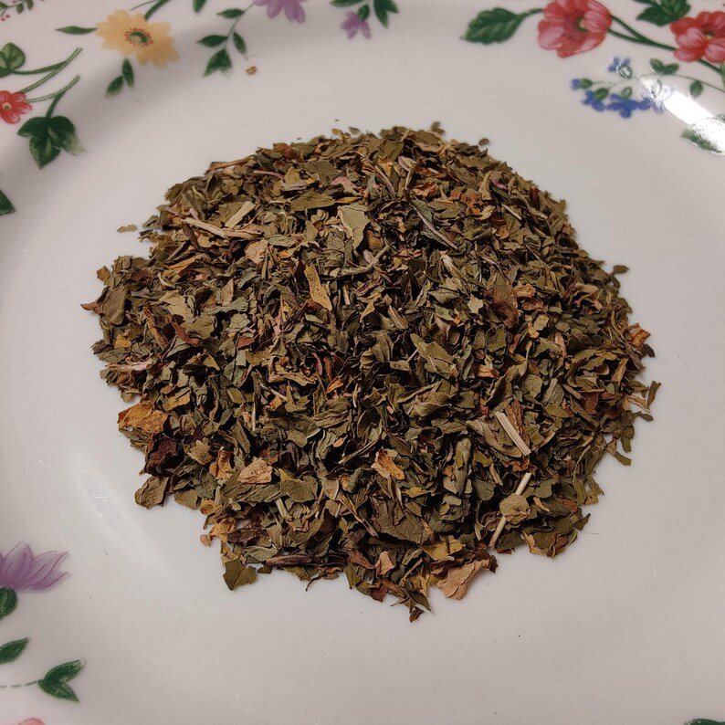 May include: A pile of dried, chopped green herbs, possibly mint or basil, on a white plate with floral designs. The herbs have a mix of green and brown hues, suggesting they are dried and ready for use in cooking or tea.