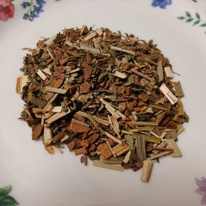 May include: A close-up shot of a pile of dried herbs and spices on a white plate. The mix includes pieces of brown bark, green leaves, and tan stalks. The plate has a floral pattern around the edge.
