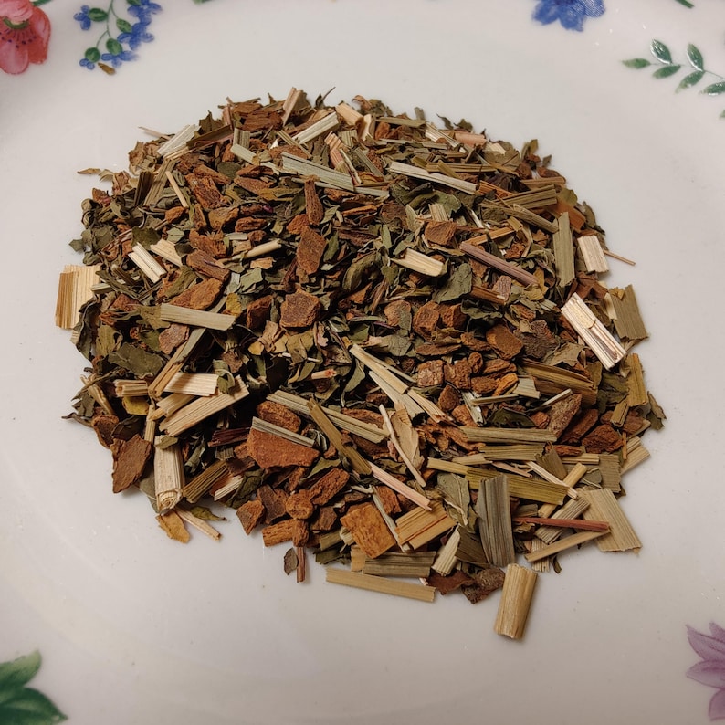 May include: A close-up shot of a pile of dried herbs and spices on a white plate. The mix includes pieces of brown bark, green leaves, and tan stalks. The plate has a floral pattern around the edge.
