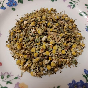 May include: A pile of dried chamomile flowers and herbs on a white plate with a floral pattern. The chamomile flowers are yellow and the herbs are green and brown. The plate has a blue, purple, and pink floral design.