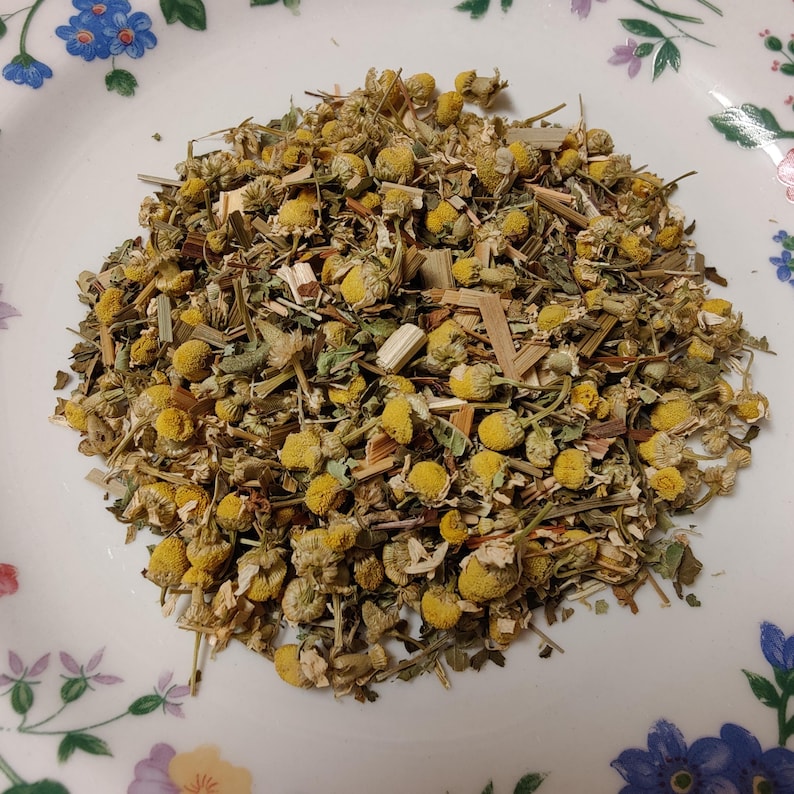 May include: A pile of dried chamomile flowers and herbs on a white plate with a floral pattern. The chamomile flowers are yellow and the herbs are green and brown. The plate has a blue, purple, and pink floral design.