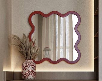 Puede incluir: Un espejo de pared con un marco rojo ondulado y festoneado. Junto al espejo hay un jarrón decorativo con un patrón geométrico rojo y blanco, lleno de hierba de la pampa seca. El espejo refleja una ventana con cortinas translúcidas.