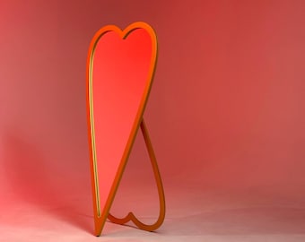 Puede incluir: Un espejo dorado en forma de corazón con una superficie reflectante roja. El espejo está de pie sobre una superficie blanca.