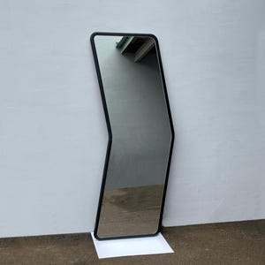 May include: A black framed full-length mirror with a unique, angled design. The mirror is standing on a white surface.