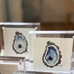 May include: Watercolor paintings of oyster shells in shades of blue and gray, displayed in clear acrylic frames. The artwork features a detailed depiction of the oyster's interior, with a signature in the bottom right corner.
