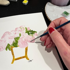 May include: Watercolor painting of pink flowers in a gold-colored vase. Green leaves are interspersed among the flowers. A hand with red nail polish holds a paintbrush, adding details to the painting. Watercolor supplies are in the background.
