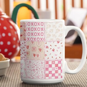 May include: White ceramic mug with a patchwork design in pink and cream. The mug features hearts, cherubs, and the text "XOXO". A red teapot with white polka dots is in the background. The mug is suitable for tea or coffee.
