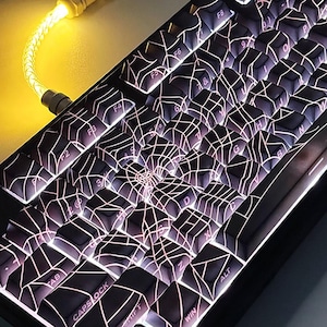 Puede incluir: Un teclado mecánico negro con un diseño de telaraña en las teclas. Las teclas son blancas con acentos rosados y brillan en la oscuridad. El teclado tiene un marco negro y una retroiluminación azul.